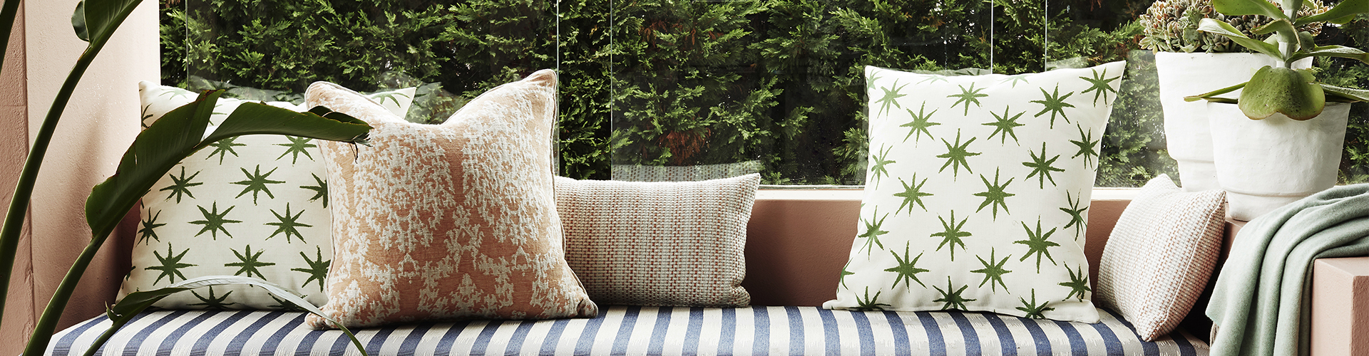 A built-in window seat layered with patterned cushions in greens, terracotta, and natural woven textures over a blue-and-white striped base, finished with a soft sage throw. Potted succulents and trailing greenery frame the nook, with lush garden views through the glass beyond.