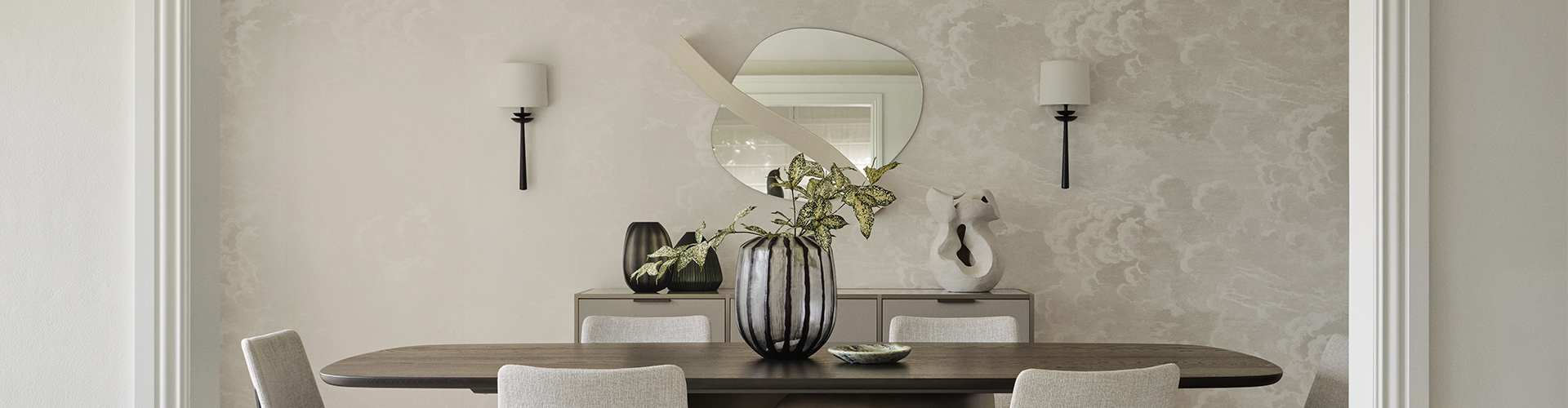 An elegant dining room centred on an oval timber table surrounded by cream upholstered chairs, set against soft patterned wallpaper. An organic-shaped mirror and twin black wall sconces sit above a sideboard styled with sculptural vases and a fluted glass vessel.